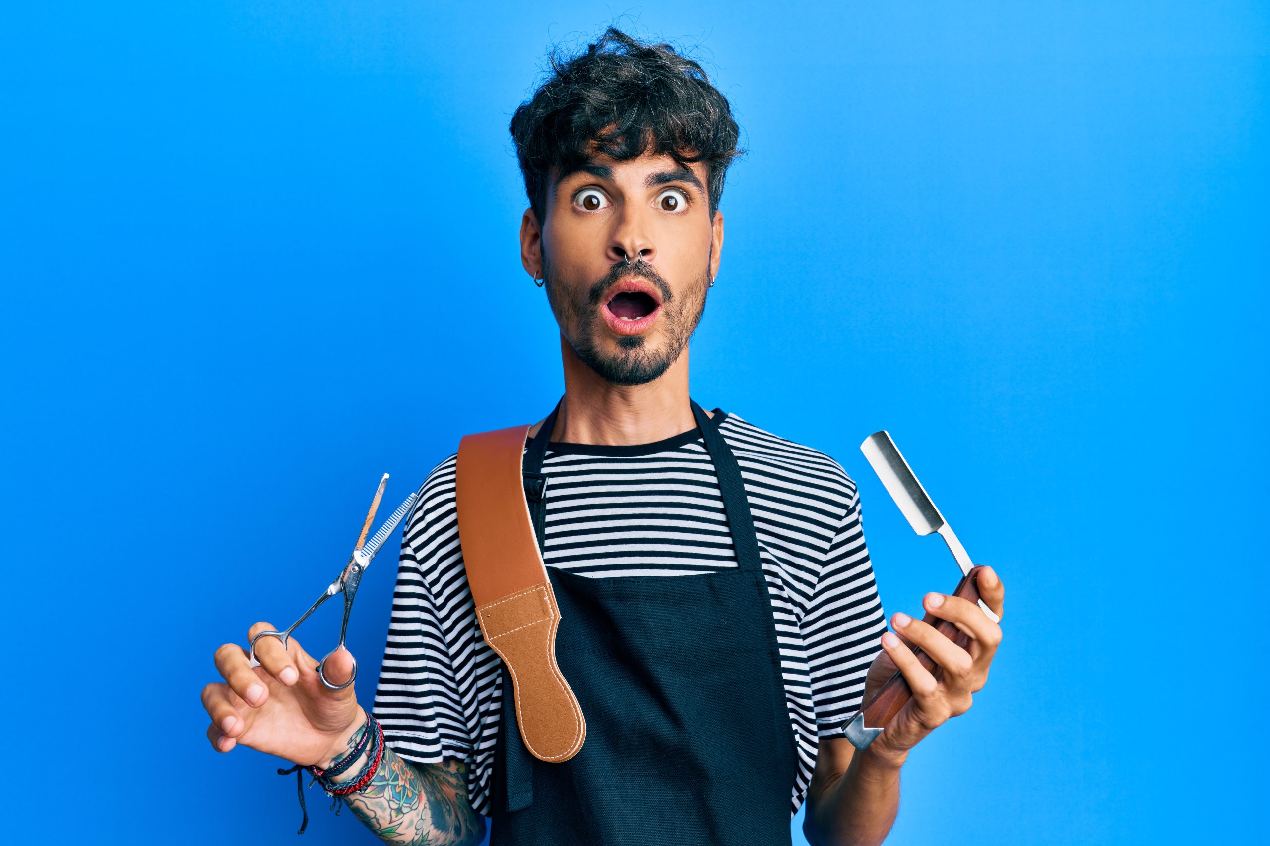 Young,Hispanic,Man,Wearing,Barber,Apron,Holding,Razor,And,Scissors
